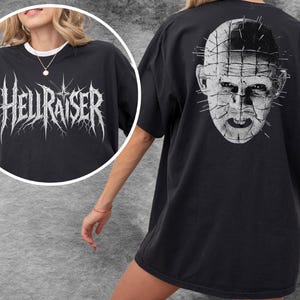 Pode incluir: Uma t-shirt preta com uma estampa gráfica branca. A frente exibe o texto HELLRAISER numa fonte estilizada e irregular. As costas apresentam uma ilustração branca detalhada da personagem Pinhead com alfinetes na cabeça e no rosto.