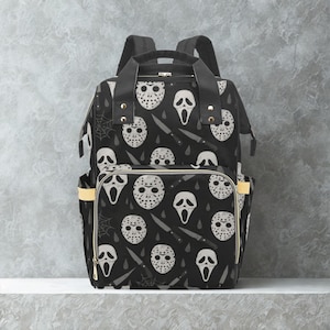 May include: Black backpack featuring a repeating pattern of white horror movie masks, knives, and spiderwebs. The bag has two side pockets with yellow accents and a top handle. The bag has two shoulder straps.