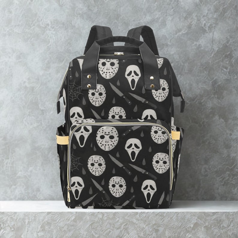 May include: Black backpack with a repeating pattern of white horror movie masks, knives, and spiderwebs. The bag has two side pockets with yellow accents and a top handle. The bag has two shoulder straps.