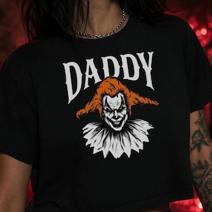 Puede incluir: Una camiseta corta negra con la palabra DADDY en letras mayúsculas blancas sobre una ilustración gráfica de un payaso con pelo naranja, cara blanca con marcas negras y un cuello de volantes blanco. El diseño está centrado en el pecho.