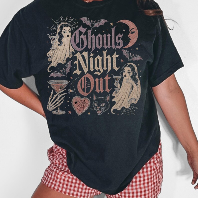 Puede incluir: Una camiseta negra de manga corta con un estampado gr&aacute;fico que incluye el texto Ghouls Night Out. El dise&ntilde;o muestra dos figuras fantasmales con copas de martini, un gato negro, murci&eacute;lagos, una luna creciente, una telara&ntilde;a y un coraz&oacute;n.