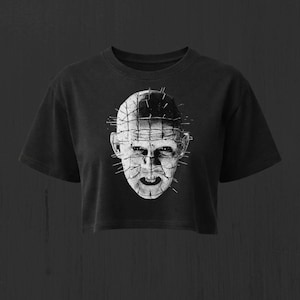 May include: A black cropped t-shirt featuring a white graphic print of a human head covered in a grid pattern with multiple pins protruding from the skin. The design is centered on the chest, showcasing a distressed, high-contrast monochromatic style.