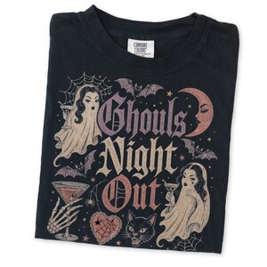 Puede incluir: Una camiseta negra con un estampado gr&aacute;fico que incluye el texto Ghouls Night Out. El dise&ntilde;o muestra dos figuras fantasmales con copas de martini, una luna creciente, murci&eacute;lagos, una telara&ntilde;a, un coraz&oacute;n y un gato negro en tonos rosa y crema.