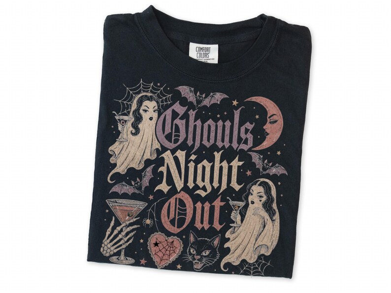 Puede incluir: Una camiseta negra con un estampado gr&aacute;fico que incluye el texto Ghouls Night Out. El dise&ntilde;o muestra dos figuras fantasmales con copas de martini, una luna creciente, murci&eacute;lagos, una telara&ntilde;a, un coraz&oacute;n y un gato negro en tonos rosa y crema.