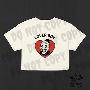 Puede incluir: Top corto color crema con un gráfico de una cara de payaso dentro de un corazón rojo. El texto "LOVER BOY" está encima del corazón. La camiseta tiene mangas cortas y cuello redondo. El logotipo de Nocturnal Nursery Co. está en la parte inferior.