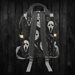 May include: Black backpack featuring a repeating pattern of white horror movie masks, knives, and spiderwebs. The backpack has a top handle, adjustable shoulder straps, and side pockets. The bag is made of a durable material.