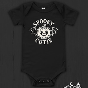 May include: Black baby bodysuit with a white graphic that says "Spooky Cutie" with a pumpkin and bat wings. The bodysuit has short sleeves and snaps at the bottom. The Nocturnal Nursery Co. logo is at the bottom right.