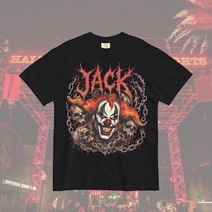 Puede incluir: Una camiseta negra de manga corta con un estampado gráfico de una cara de payaso con pelo naranja, maquillaje verde en los ojos y nariz roja. El diseño incluye la palabra JACK en una fuente roja dentada, rodeada de calaveras y cadenas.
