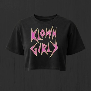 Puede incluir: Un top corto negro de manga corta con el texto KLOWN GIRLY impreso en una fuente estilizada y dentada. Las letras son de color rosa vibrante con un contorno amarillo claro, creando una apariencia nítida y angular sobre la tela oscura.