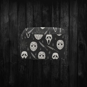 May include: Black accessory case featuring a repeating pattern of white horror movie masks, knives, and spiderwebs. The case has a rectangular shape and a black background. The design is a spooky, Halloween-themed pattern.