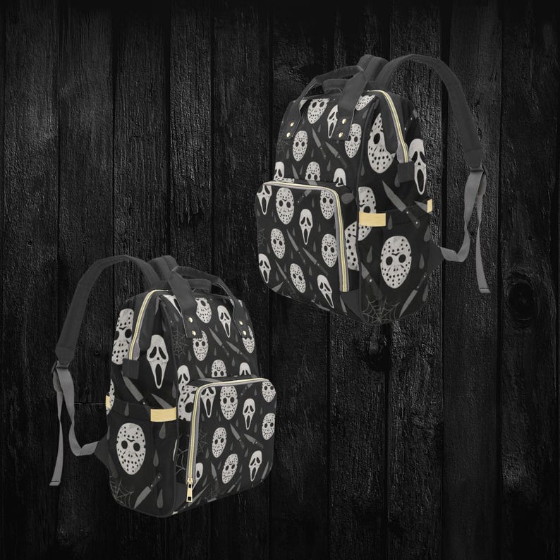 May include: Two black backpacks with a repeating pattern of white horror movie masks, knives, and spiderwebs. The backpacks have black straps, gold zippers, and a front pocket. The background is a dark wood.
