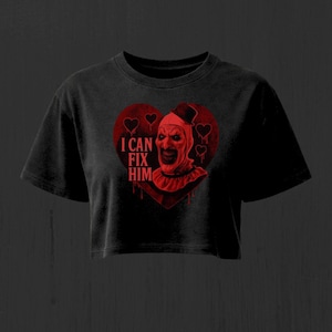 Puede incluir: Una camiseta corta negra con un gráfico rojo de un payaso con sombrero de copa dentro de un corazón. El texto "I CAN FIX HIM" está impreso en rojo a la izquierda del payaso. El diseño incluye contornos de corazones y efectos de goteo.