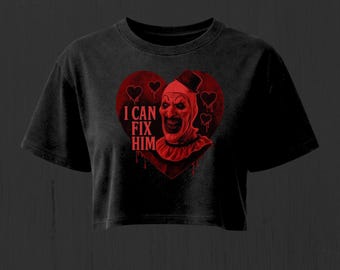 Puede incluir: Una camiseta corta negra con un gráfico rojo de un payaso con sombrero de copa dentro de un corazón. El texto "I CAN FIX HIM" está impreso en rojo a la izquierda del payaso. El diseño incluye contornos de corazones y efectos de goteo.