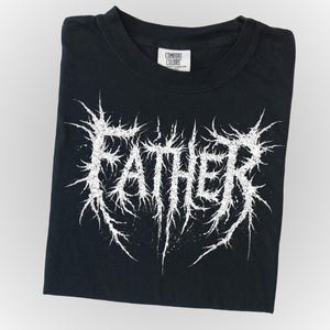 May include: Black t-shirt with the word "FATHER" in a white, spiky font. The shirt is folded neatly, with the Comfort Colors tag visible. The design has a heavy metal aesthetic.