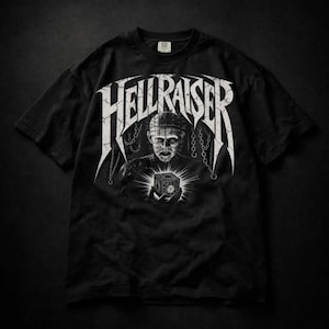 May include: A black short-sleeve t-shirt featuring a white graphic print of the character Pinhead from Hellraiser holding a puzzle box. The word HELLRAISER is printed in a large, distressed white font above the character, surrounded by hanging chains.