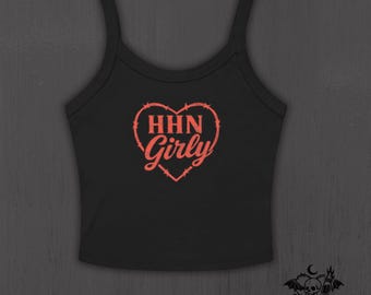 May include: A black sleeveless crop top featuring a coral-colored graphic of a heart formed by barbed wire. Inside the heart, the text reads HHN Girly in a bold, stylized font. The garment has a scoop neckline and wide shoulder straps.