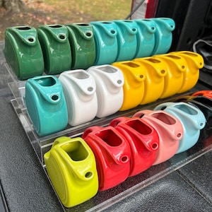 May include: A collection of colorful ceramic creamer pitchers in various shades of green, blue, yellow, red, and white. The pitchers are arranged in rows on a clear acrylic display stand.