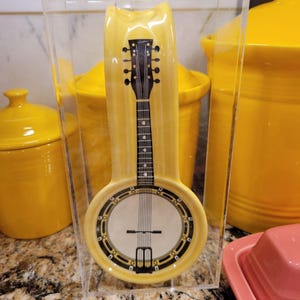 May include: A yellow ceramic banjo-shaped spoon rest inside a clear acrylic holder. The banjo design features a black neck and strings, with a white circular face and black and gold accents. Other yellow kitchenware is in the background.