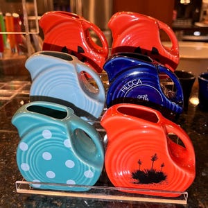 May include: A collection of six ceramic pitchers in various colors including red, light blue, dark blue, and teal. Some pitchers feature white polka dots, black desert plant silhouettes, or the text HECCA 25th. Each has a handle and spout.