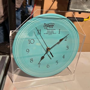 May include: A turquoise Fiestaware clock with black numbers and hands, encased in a clear acrylic stand. The clock face features the text "Genuine Fiesta 60th Anniversary 1936-1996 The Homer Laughlin China Co."