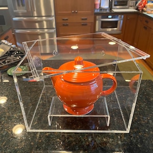 May include: An orange ceramic teapot with a matching lid, displayed inside a clear acrylic display case. The teapot sits on a clear acrylic stand. The case has a hinged lid and is on a dark countertop.