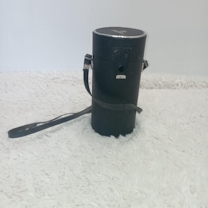 Peut inclure: Un étui cylindrique noir avec un bord argenté et une sangle en cuir noir. L'étui a une fermeture à boucle et un petit emblème sur le dessus. Il est probablement destiné à ranger un objectif d'appareil photo ou autre équipement, environ 25 cm de haut.