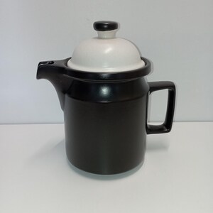 Könnte beinhalten: Schwarze Keramik-Teekanne mit weißem Deckel und schwarzem Griff. Der Deckel hat einen kleinen schwarzen Knopf. Die Teekanne hat einen Ausguss zum Eingießen von Tee. Die Teekanne steht auf einer weißen Oberfläche.