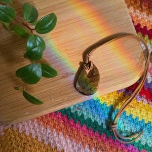 Puede incluir: Un collar con colgante de piedra natural y cordón de cuero marrón. El colgante tiene tonos verdes y marrones. El collar descansa sobre una superficie de madera con una planta y un textil colorido y estampado en el fondo. Un arcoíris de luz brilla a través de la escena.