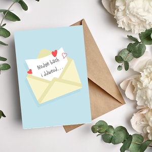 May include: A light blue greeting card featuring a yellow envelope illustration with a letter and red hearts. The letter reads "Nodyn bach i ddweud..." The card is placed next to a brown envelope and white flowers.