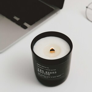 May include: A black candle with a white wax top and a gold wick. The candle is labeled "Sweet Hustle Co. Coconut | Cocoa Butter Two Weeks Notice Soy Candle Made in New Jersey"