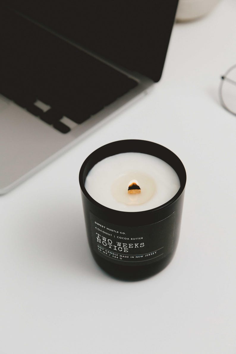 May include: A black candle with a white wax top and a gold wick. The candle is labeled "Sweet Hustle Co. Coconut | Cocoa Butter Two Weeks Notice Soy Candle Made in New Jersey"