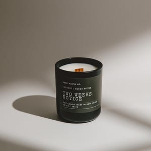 May include: A black candle jar with a wooden wick and a label that reads "Sweet Hustle Co. Coconut | Cocoa Butter Two Weeks Notice 10 oz Candle Made in New Jersey".