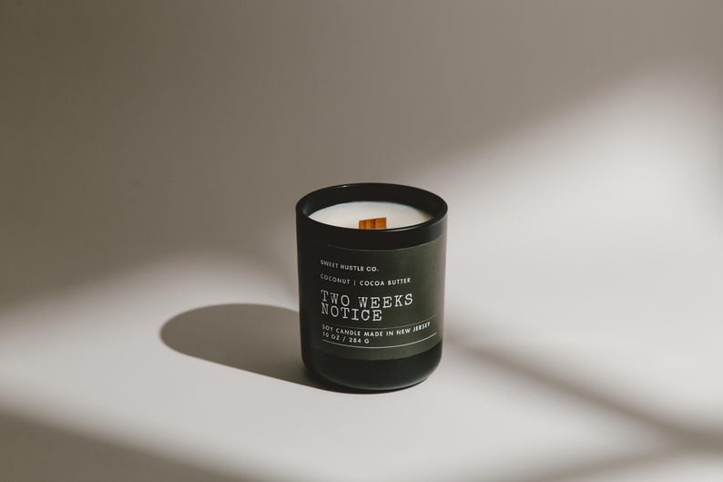 May include: A black candle jar with a wooden wick and a label that reads "Sweet Hustle Co. Coconut | Cocoa Butter Two Weeks Notice 10 oz Candle Made in New Jersey".