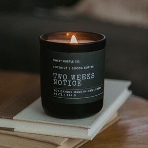 May include: A black soy candle with a white label that reads "SWEET HUSTLE CO. COCONUT | COCOA BUTTER TWO WEEKS NOTICE SOY CANDLE MADE IN NEW JERSEY 10 OZ / 284 G". The candle is lit and the flame is visible.