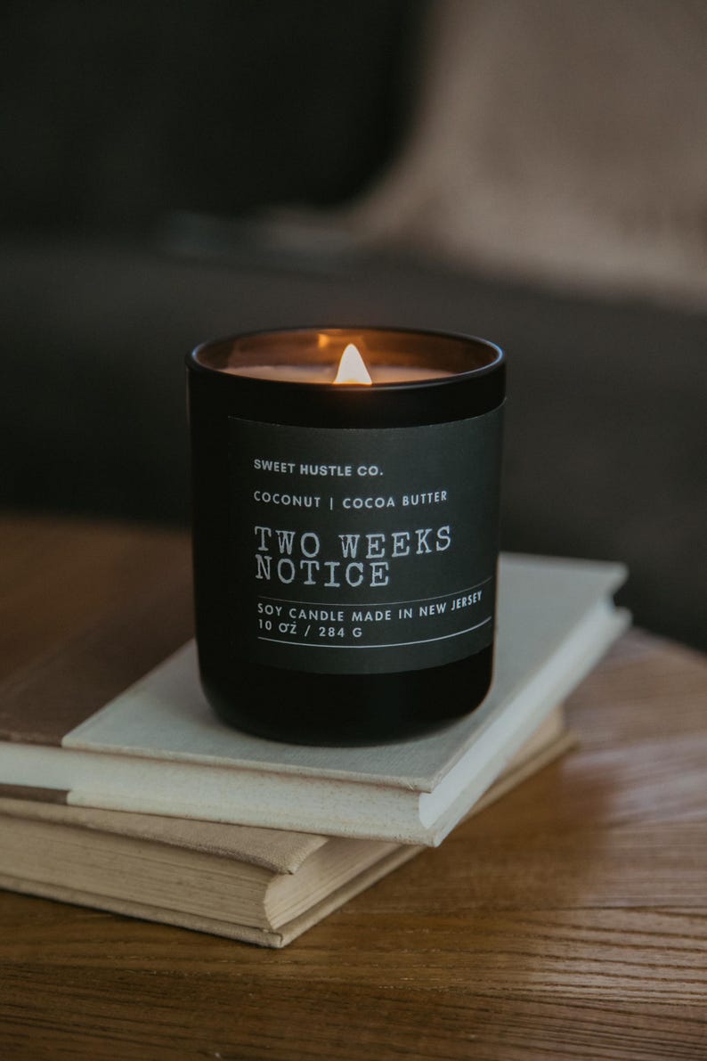 May include: A black soy candle with a white label that reads "SWEET HUSTLE CO. COCONUT | COCOA BUTTER TWO WEEKS NOTICE SOY CANDLE MADE IN NEW JERSEY 10 OZ / 284 G". The candle is lit and the flame is visible.
