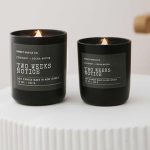 May include: Two black soy candles with white labels. The labels read "Sweet Hustle Co. Coconut | Cocoa Butter Two Weeks Notice Soy Candle Made in New Jersey 10 oz / 284 g".