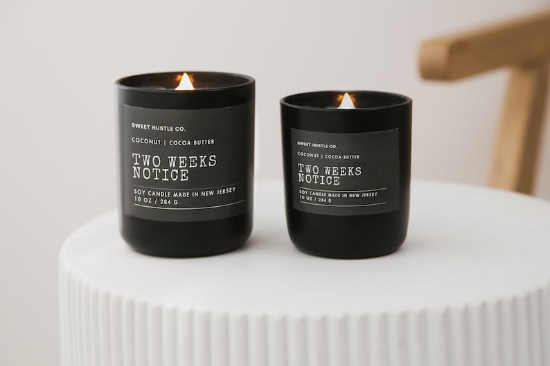 May include: Two black soy candles with white labels. The labels read "Sweet Hustle Co. Coconut | Cocoa Butter Two Weeks Notice Soy Candle Made in New Jersey 10 oz / 284 g".