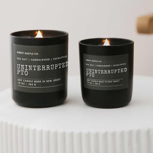 May include: Two black soy candles with white labels. The labels read "Sweet Hustle Co." and "Uninterrupted PTO." The candles are lit and have a warm glow.