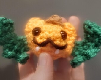 Op de afbeelding: Een gehaakte pompoen met een groene vleermuisvleugel aan elke kant. De pompoen heeft een bruine snor en zwarte ogen.