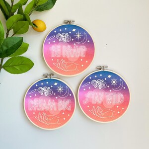 Könnte beinhalten: Drei Stickrahmen mit einem rosa-violetten Farbverlauf als Hintergrund. Jeder Rahmen zeigt eine weiße Umrandung einer Hand, die eine himmlische Szene mit Sternen, Monden und Planeten hält. Der Text auf den Rahmen lautet "Be Brave", "Dream Big" und "Stay Wild".