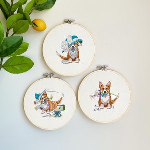 Op de afbeelding: Drie ronde borduurringen met corgi-hondontwerpen. Eén corgi draagt een hoed en parels, een andere heeft papieren en een kopje thee, en de derde houdt bloemen vast. De ringen staan tegen een witte achtergrond.