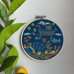 Könnte beinhalten: Ein blauer Stickrahmen mit einem farbenfrohen Design aus einem Boot, Fischen, Algen und einem Seestern. Das Design befindet sich auf einem dunkelblauen Hintergrund mit weißen Sternen.