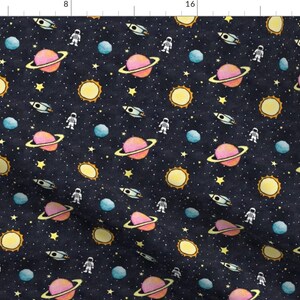 May include: A dark blue fabric with a repeating pattern of planets, stars, astronauts, and rockets. The planets are pink, yellow, and blue. The stars are yellow. The astronauts are white. The rockets are gray.