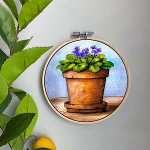 May include: A hand-painted embroidery hoop featuring a potted violet plant with purple flowers and green leaves. The plant is in a brown terracotta pot with a light blue background.