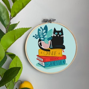 Peut inclure: Un chat noir assis sur une pile de livres avec une tasse de thé. Les livres sont rouge, jaune et bleu. Le texte sur les livres est "One more chapter".