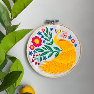 May include: A yellow peacock with a pink butterfly embroidered on a white fabric stretched over a wooden embroidery hoop. The peacock is surrounded by green leaves and pink, blue, and red flowers.