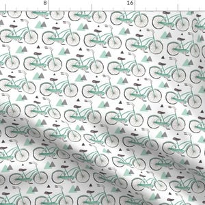 Op de afbeelding: Witte stof met een herhalend patroon van turquoise fietsen en grijze driehoeken. De fietsen zijn getekend met zwarte contouren en hebben zwarte zadels en sturen. De driehoeken zijn in verschillende maten en oriëntaties.