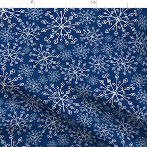 Op de afbeelding: Een donkerblauwe stof met een herhalend patroon van witte sneeuwvlokken. De sneeuwvlokken variëren in grootte en hebben gedetailleerde, sierlijke ontwerpen. Geschikt voor winterthema projecten of kleding.