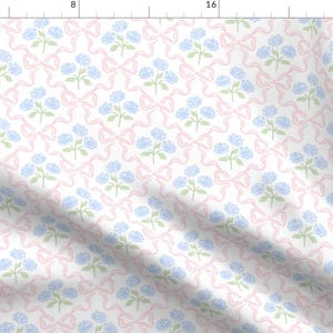 以下が含まれることがあります： ピンクのリボンと青い花が繰り返し描かれた白い生地。繊細で女性らしいパターンです。