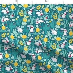 May include: A teal blue fabric with a repeating pattern of white rabbits, yellow sunflowers, green cacti, and yellow turtles. The rabbits are wearing red scarves.